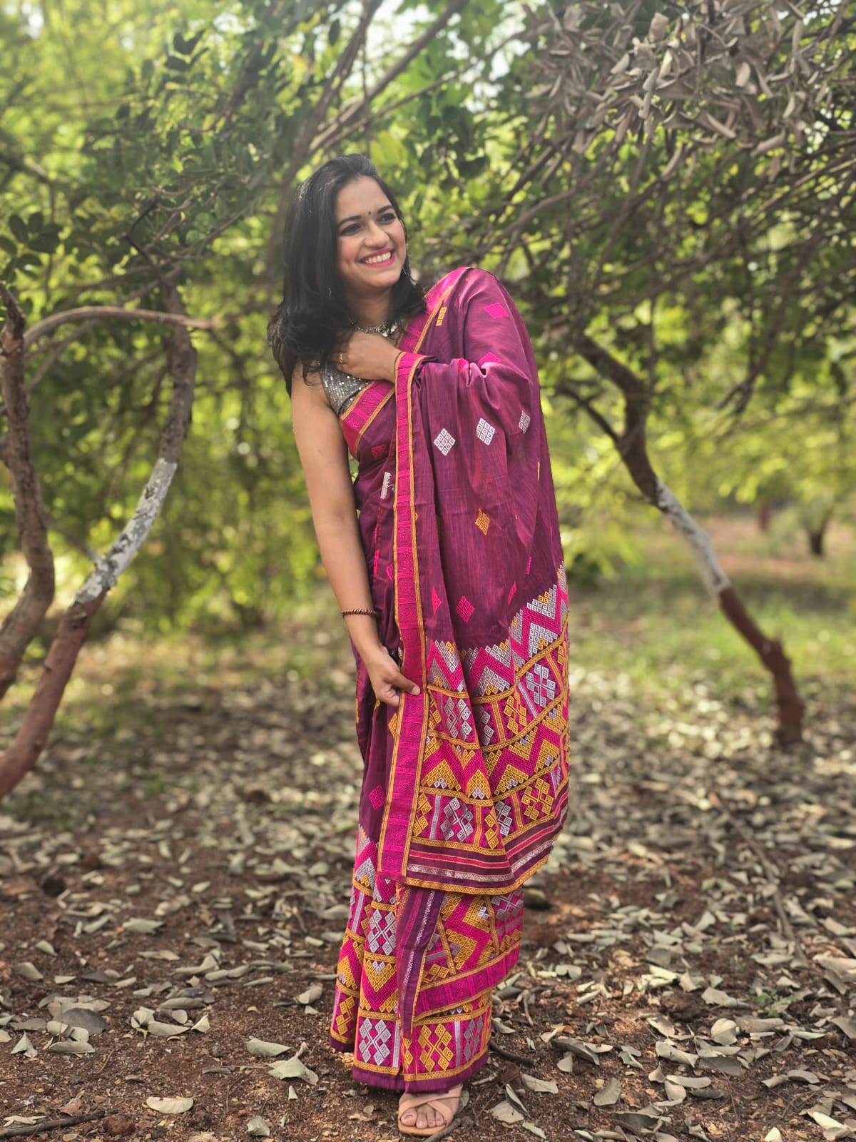 A woman smiling while draping The Dawn Ligang, a vibrant pink handwoven Mishing Mekhela Chador with intricate yellow and magenta geometric motifs.